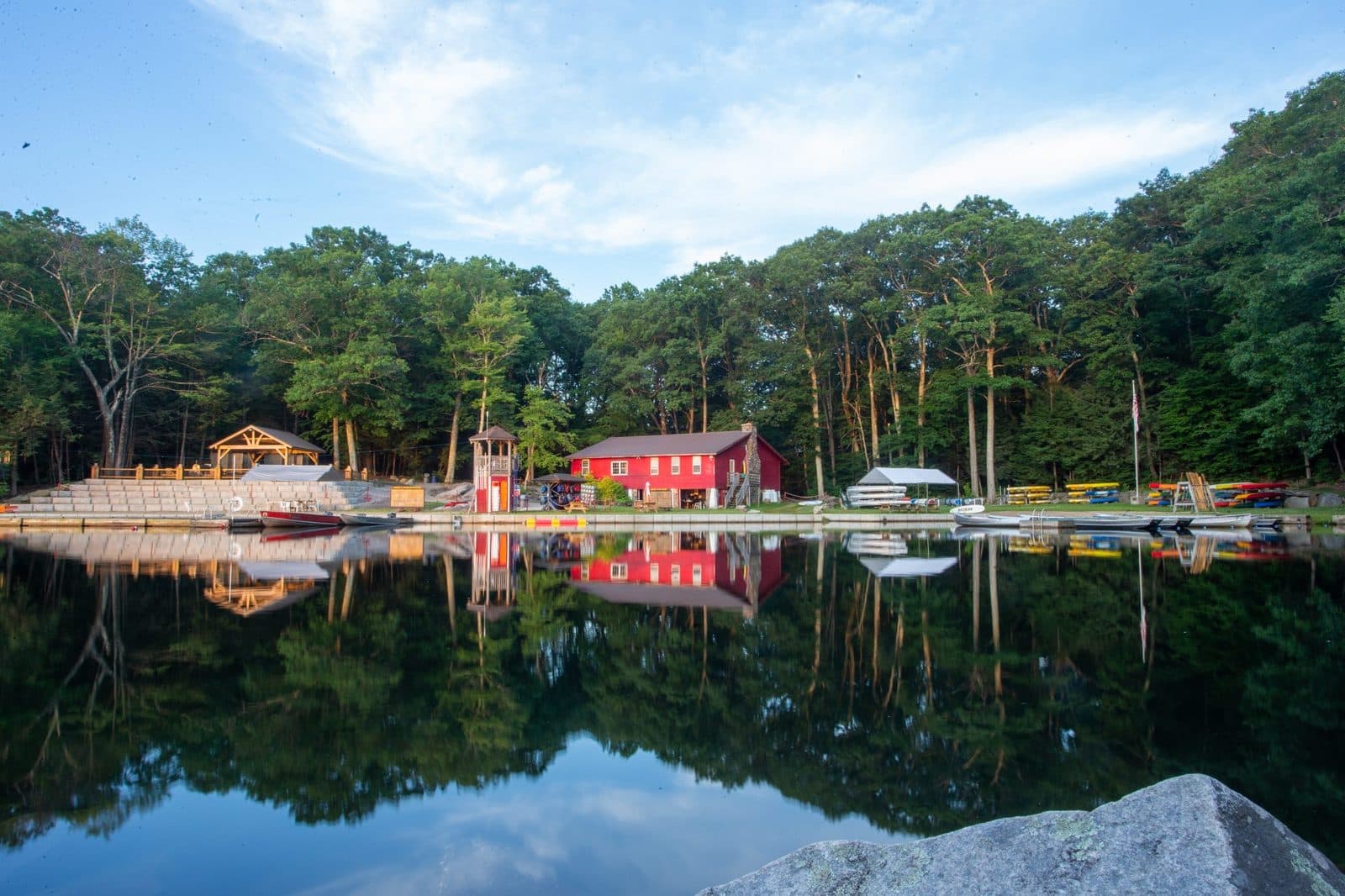 The lake at Camp Sequassen on a still morning, its red waterfront buildings and the treeline mirrored in the water
